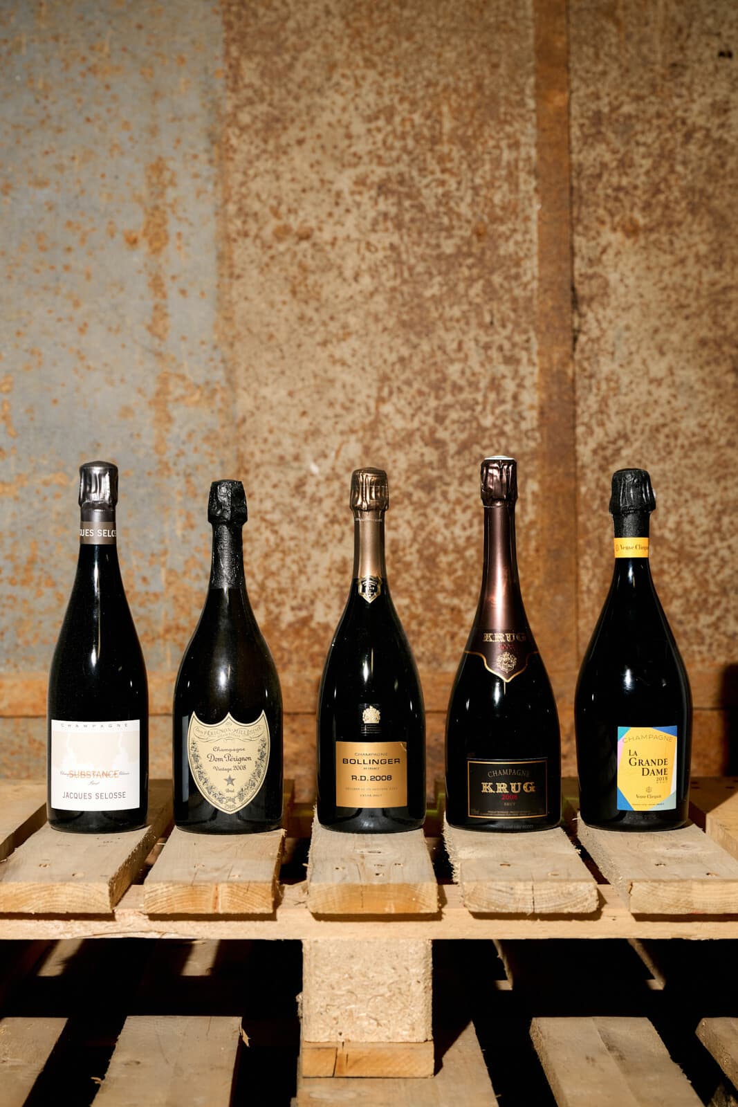 Pět lahví šampaňského na paletě před rezavou stěnou: Jacques Selosse, Dom Pérignon, Bollinger R.D., Krug a La Grande Dame.