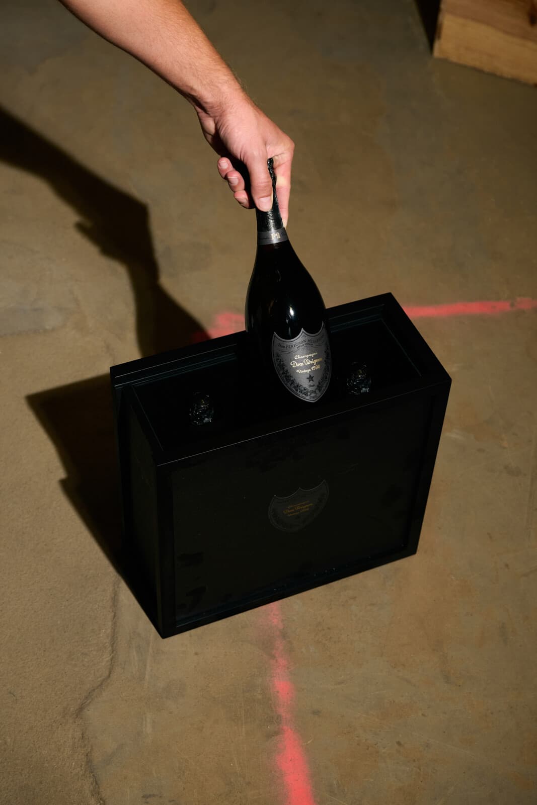 Ruka vkládá lahev Dom Pérignon P2 Vintage 1998 do černé dárkové kazety na betonové podlaze.