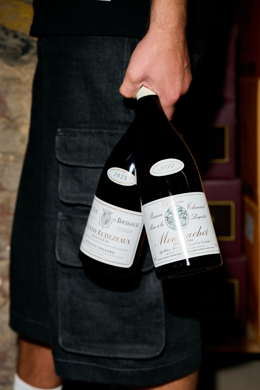 Ruka nese za hrdla dvě lahve Domaine Thénard: Grands-Échezeaux Grand Cru a Montrachet Grand Cru.