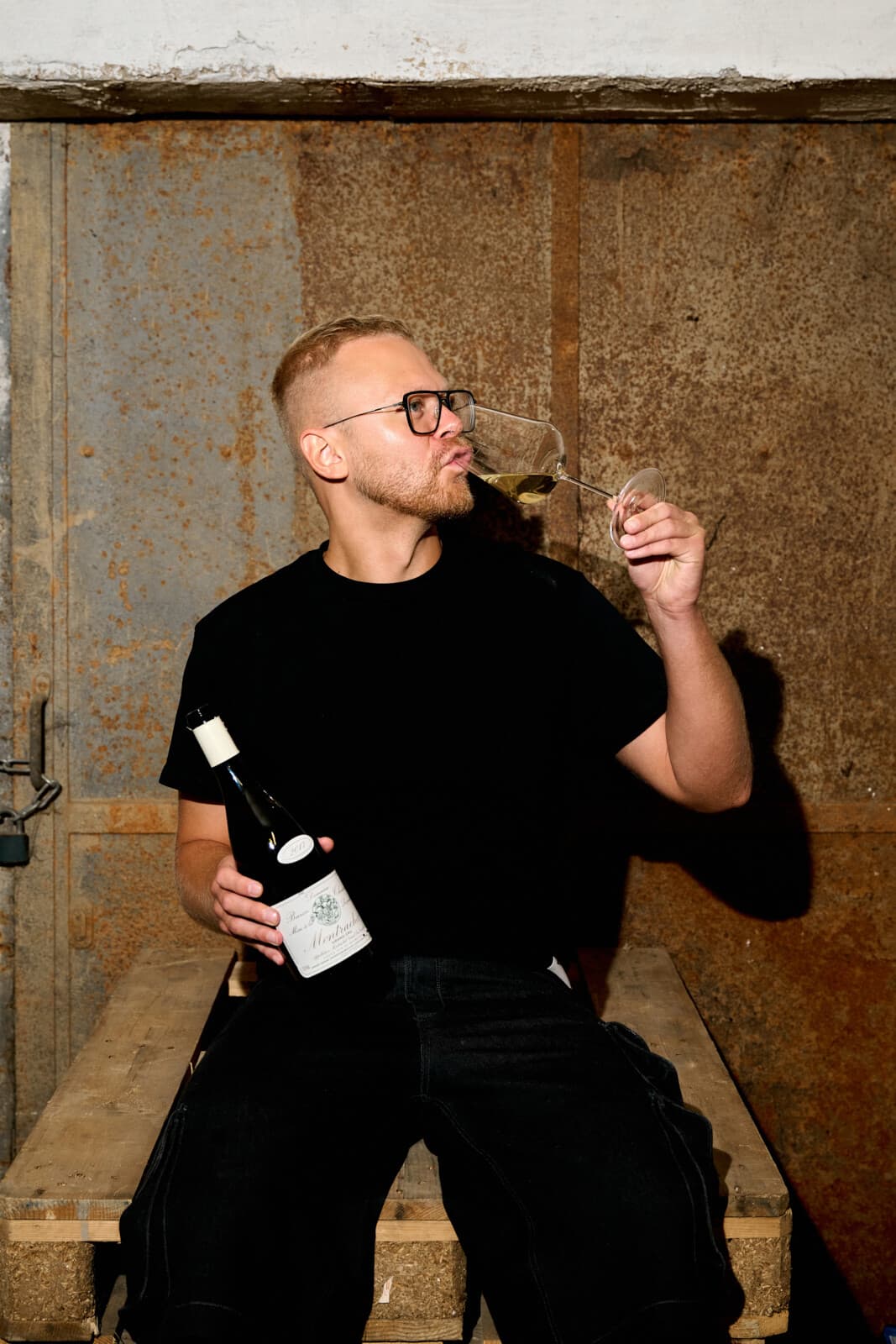 Jakub Šebela ochutnává bílé víno, v druhé ruce drží lahev; sedí na paletě před rezavými dveřmi sklepa.