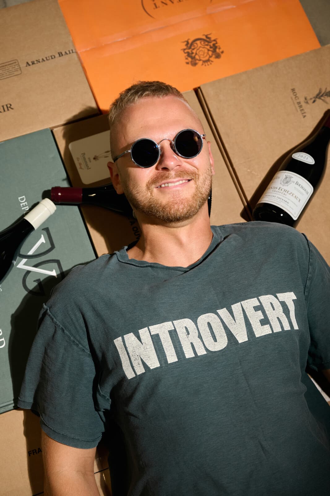 Jakub Šebela v kulatých slunečních brýlích leží na kartonech vín, kolem něj lahve.