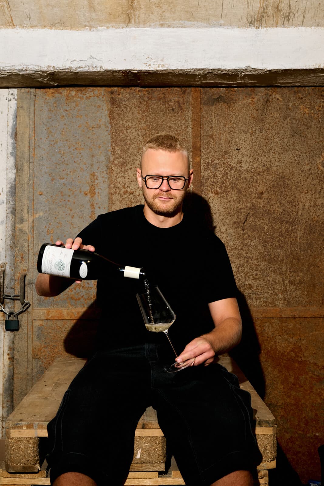 Jakub Šebela nalévá bílé víno do sklenky, sedí na paletě před rezavými dveřmi sklepa.