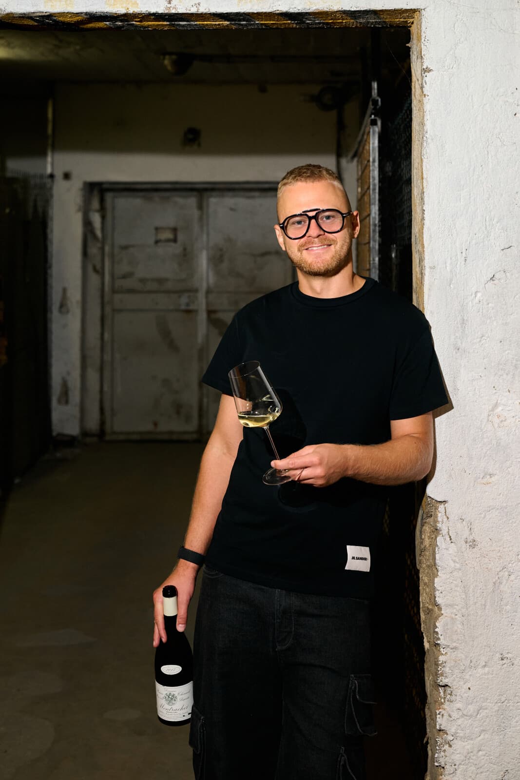 Jakub Šebela opřený o bílou zeď ve dveřích sklepa, se sklenkou vína a lahví v ruce.